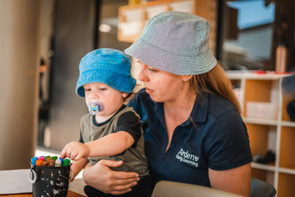 An Arden educator cuddling a toddler in a blue bucket hat