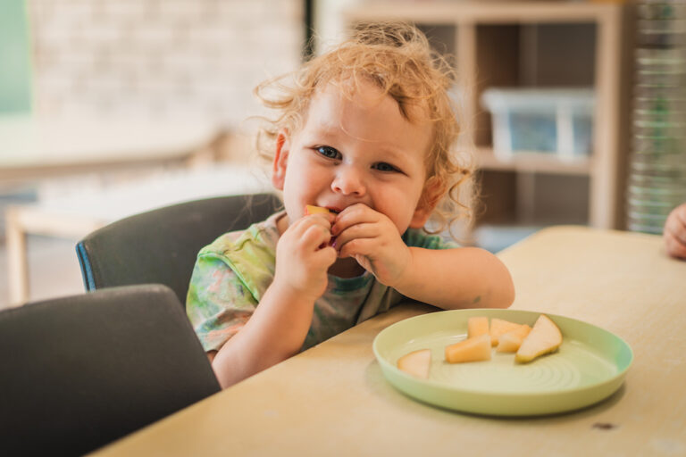 A smiling toddler enjoying a snack of fruit at the table