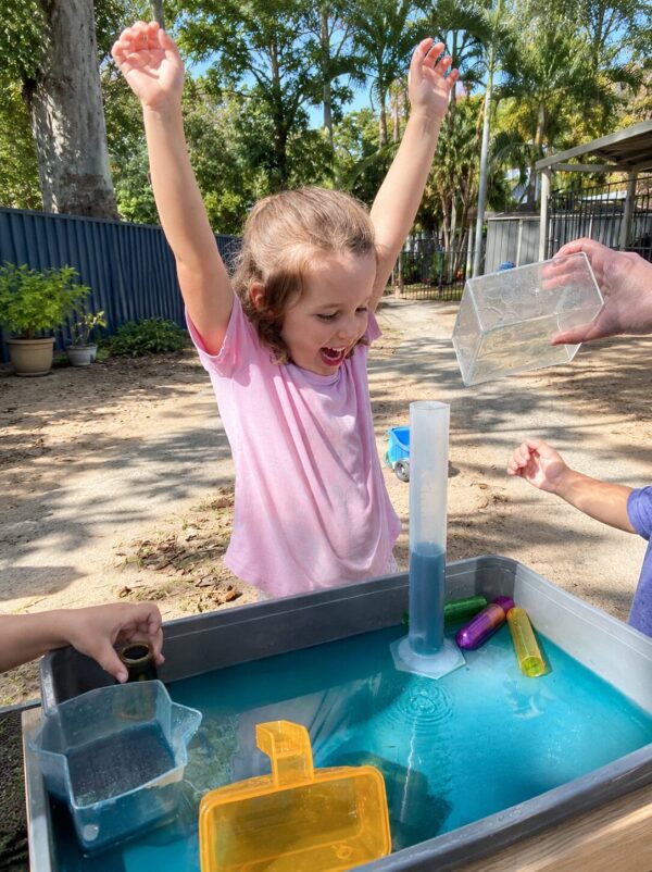 A child celebrating with arms raised at the outdoor water play table at Arden Early Learning Airlie Beach